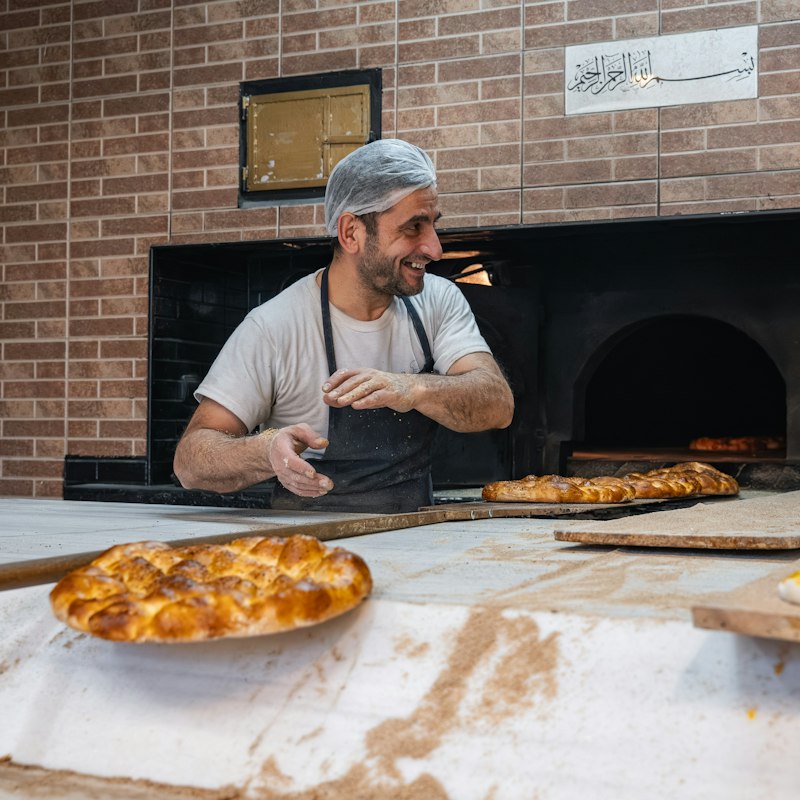 Baker smiling beside a brick oven with fresh baked bread